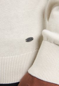 Pulover tricotat de culoare crem, cu manșete și tiv cu nervuri. Prezintă un mic tag metalic cu logo-ul "Petrol". Textură netedă, design minimalist.