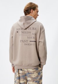 Sudadera beis con un diseño de texto grande en la parte posterior. Cuenta con una capucha ajustable y puños acanalados. Se combina con pantalones de camuflaje.