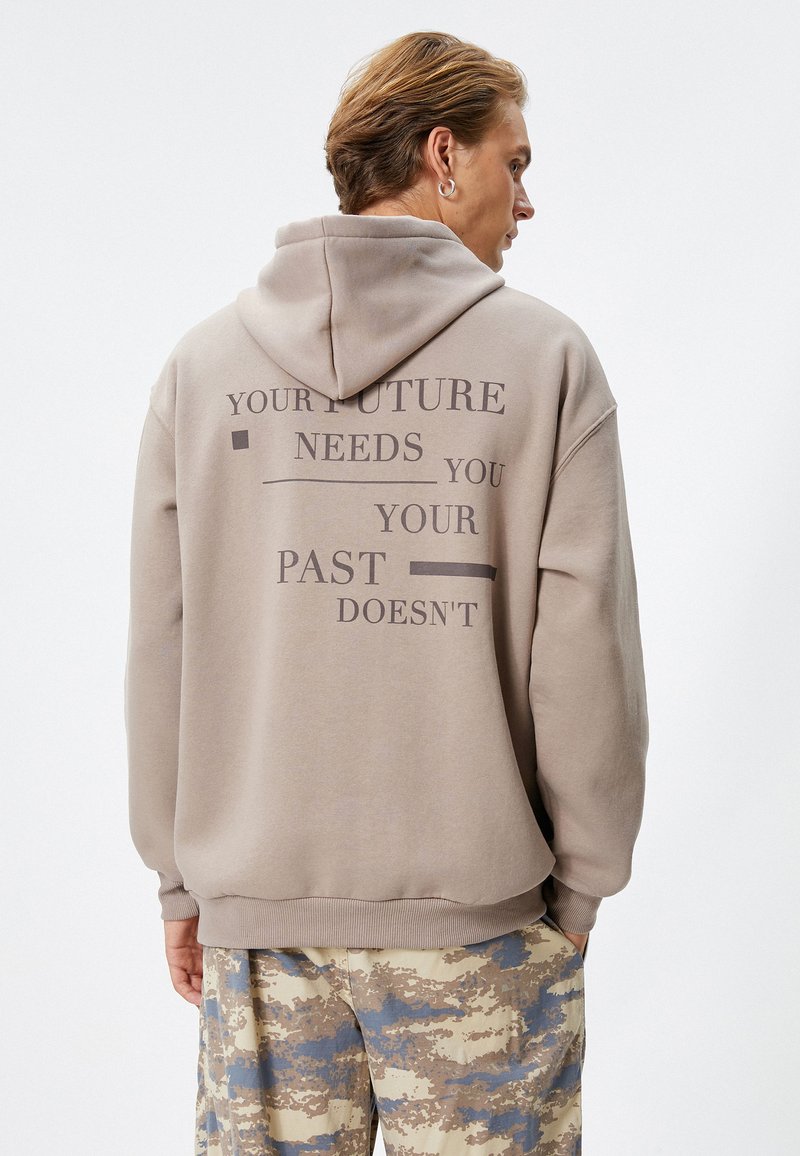 Sudadera beis con un diseño de texto grande en la parte posterior. Cuenta con una capucha ajustable y puños acanalados. Se combina con pantalones de camuflaje.