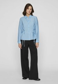 Camisa de botón a rayas azul claro con detalles atados en los laterales, combinada con pantalones anchos negros. Textura de tela suave, ajuste casual, mangas largas.