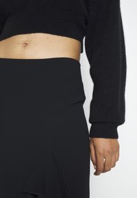 Gros plan sur la taille d’une personne portant un pull court noir et une jupe noire taille haute avec un détail volanté à l’avant.