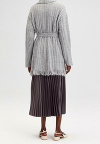 Šedý žebrovaný kardigan s páskem v pase, třásněmi na lemu, v kombinaci s plisovanou tmavě šedou sukní a krémovými sandály na širokém podpatku.