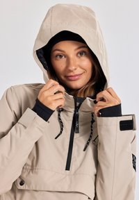 Beige waterdichte jas met een capuchon, zwarte binnenkant, verstelbare trekkoorden, voorzak en zwarte accenten bij de manchetten en de kraag.