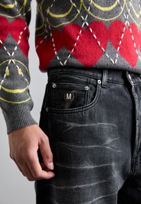 Gray sweater with red and yellow patterns, paired with black jeans featuring a faded texture and a circular brand label on the front pocket.