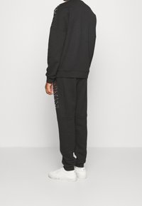 Persona che indossa una felpa nera con cappuccio e pantaloni della tuta abbinati con loghi laterali, abbinati a sneaker bianche, in piedi contro uno sfondo semplice.