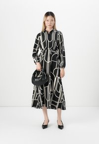 Femme portant une robe midi à manches longues avec un motif géométrique noir et blanc, tenant un sac à main noir et portant des chaussures pointues noires.