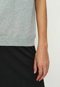 Pulover tricotat gri cu guler rotund, având un tiv cu model în relief. Asortat cu o fustă neagră cu model în relief, arătând o mână sprijinită pe lateral.