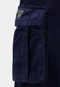 Tessuto a coste color navy con tasca a toppa e etichetta con logo. Presenta una texture a righe verticali e un lembo pieghevole sulla tasca per la chiusura.