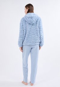 Mujer de pie descalza, vistiendo una sudadera de felpa azul claro con estampado de corazones y pantalones de pijama a lunares a juego, de espaldas contra un fondo blanco.