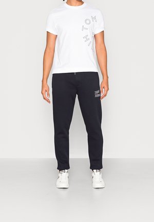 Hvid kortærmet t-shirt med sort grafisk tekst, parret med marineblå joggers med logo-tryk på venstre lår og elastiske manchetter. Hvide sneakers.
