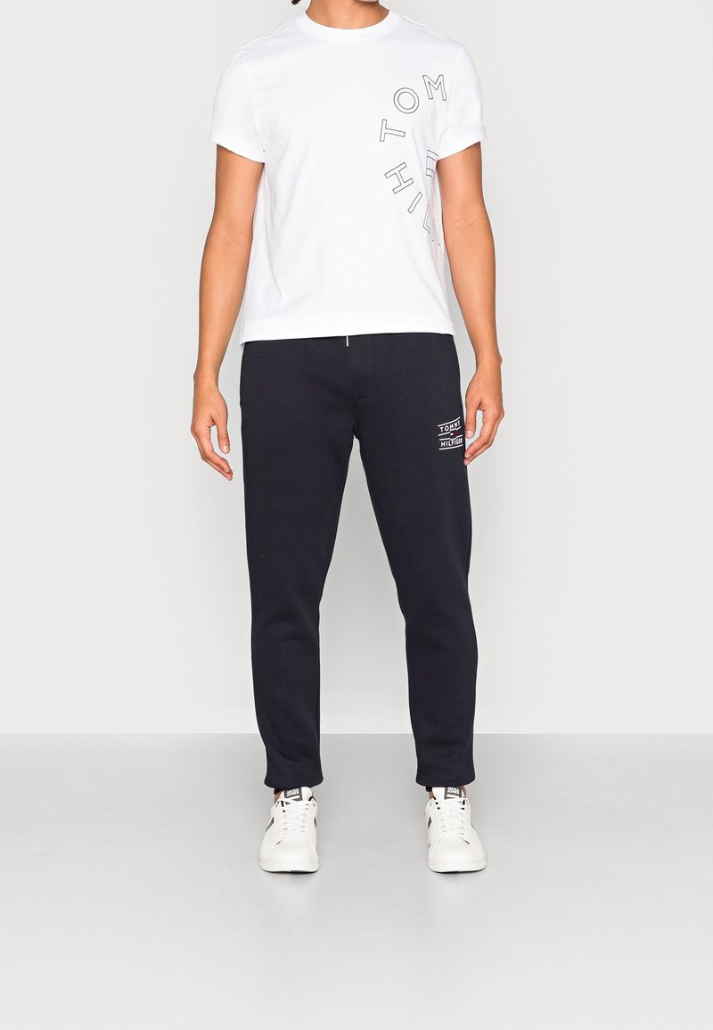 Witte t-shirt met korte mouwen en zwarte grafische tekst, gecombineerd met navy joggers met een logo-opdruk op de linkerdij en elastische boorden. Witte sneakers.