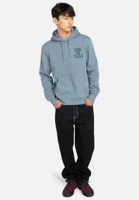 Felpa azzurra chiara con tasca frontale e testo stampato, abbinata a pantaloni neri e sneakers rosso scuro. Tessuto morbido, vestibilità casual.