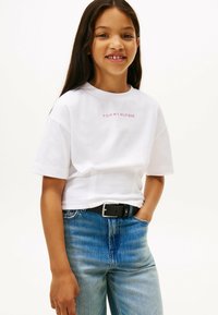 Biała bawełniana koszulka z krótkim rękawem, z napisem "TOMMY HILFIGER" na wysokości klatki piersiowej w różowym kolorze. W komplecie z niebieskimi jeansami i czarnym paskiem.