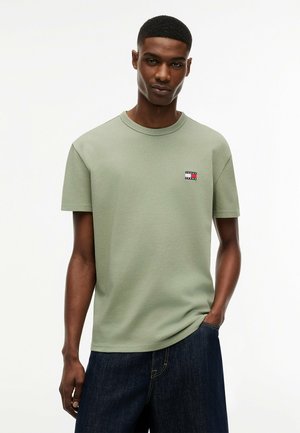 Ung mand iført en lysegrøn kortærmet T-shirt med et lille logo på brystet og mørkeblå jeans, stående med den ene hånd i lommen.