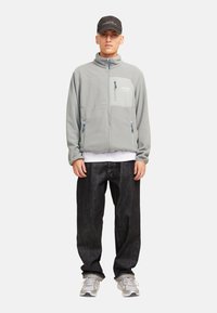 Chaqueta de forro polar gris con cuello alto, cremallera frontal, un bolsillo en el pecho y acentos en azul en las cremalleras. Lleva puesta con jeans oscuros y zapatillas grises.