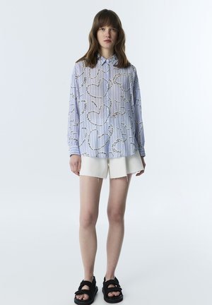 Jeune femme aux longs cheveux bruns portant une chemise bleue à motifs avec des boutons, un short blanc et des sandales noires, debout devant un fond uni.