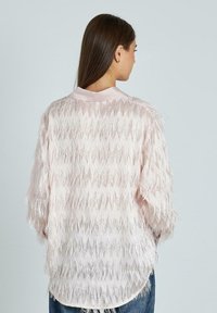 Vrouw met lang bruin haar die met de rug naar de camera staat, gekleed in een lichtroze blouse met textuur en blauwe jeans tegen een effen achtergrond.