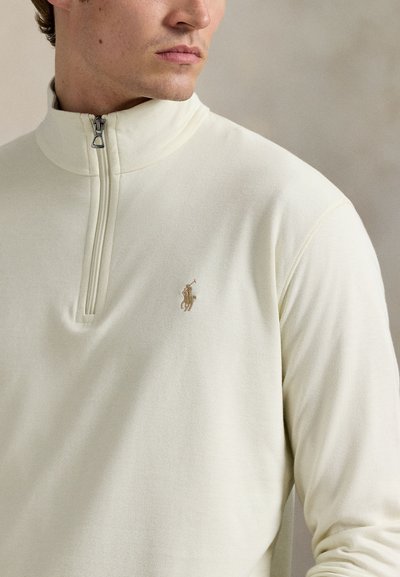 Bărbat purtând pulover crem cu fermoar de un sfert și logo brodat cu jucător de polo pe pieptul stâng, privind în lateral.