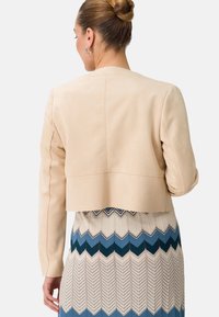 Chaqueta beige recortada con textura suave, que presenta mangas largas y corte recto. Llevada sobre una falda estampada en azul y crema.