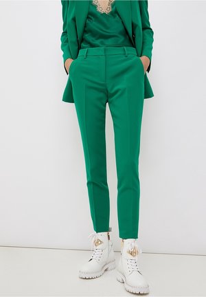 Person iført grønne skræddersyede bukser, matchende grøn blazer og hvide snørebåndsstøvler med chunky såler og guld lynlåsdetaljer.
