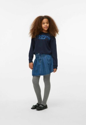 Fille aux cheveux bouclés portant un sweat à capuche bleu marine, une jupe en denim plissée bleue, des collants gris et des chaussures noires, se tenant devant un fond blanc.