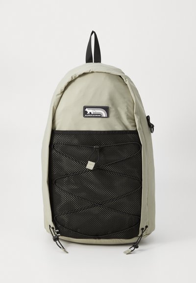 Mochila gris claro con una parte superior redondeada, que cuenta con un panel frontal de malla negra y un acento de cordón. Incluye un asa superior y un parche con el logo.