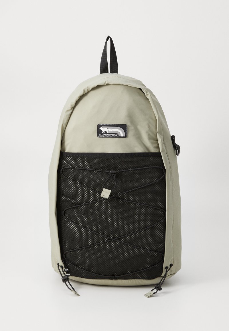 Mochila cinza-claro com uma parte superior arredondada, apresentando um painel frontal de malha preta e um detalhe em cordão. Inclui uma alça superior e um emblema.