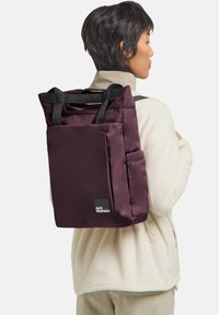 Maroon backpack with two black handles, zip closure, side pocket, and branded tag. Made from smooth fabric; rectangular shape.
