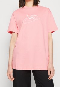 Růžové bavlněné tričko s krátkými rukávy, kulatým výstřihem a bílým vyšitým logem Nike na hrudi. Volný střih a hladká textura.
