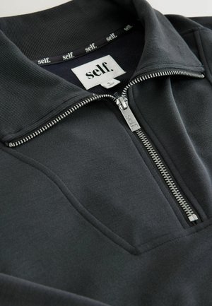 Melnā zip-up puloveris ar stāvošu apkakli, izgatavots no mīksta, teksturēta materiāla. Satur sudraba rāvējslēdzēju un etiķeti iekšpusē kaklā.