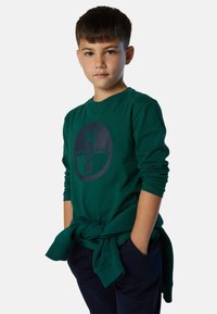 Camisa de manga larga verde con un logo gráfico redondo en color navy y texto, combinada con pantalones navy y una camisa atada a la cintura.