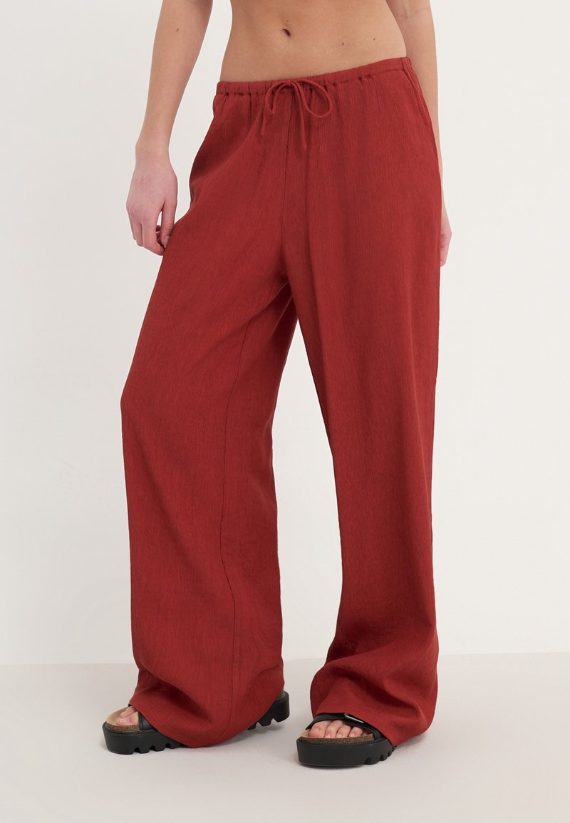 Pantalon ample, taille haute, rouge avec cordon, porté avec des sandales à plateforme noires sur une personne debout sur un fond uni.
