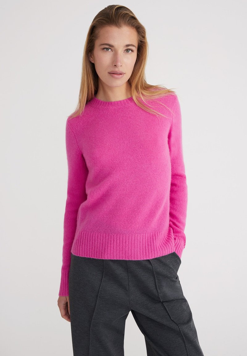 Maglione in cashmere rosa acceso con scollo rotondo, polsini e orlo a coste. Texture morbida, silhouette aderente, abbinato a pantaloni grigi scuri.