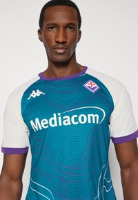 Turkizna športna majica z belimi rokavi, z logotipom 'Mediacom', vijoličnimi poudarki in valovitim vzorcem. Grb ekipe na prsih.