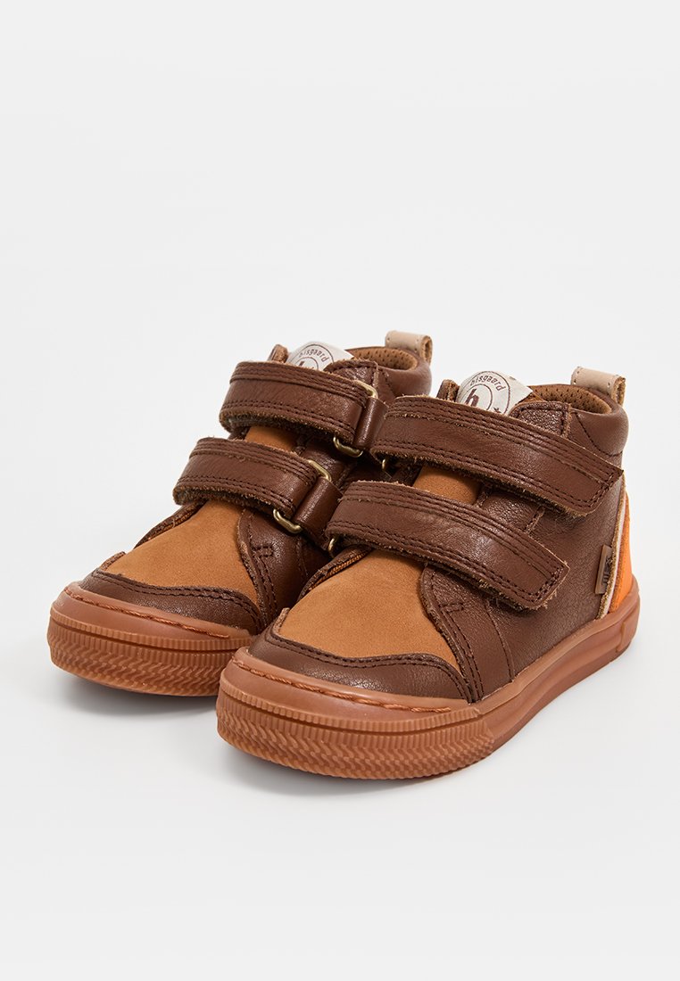 Bruine leren en beige suède kinderschoenen met twee klittenbandsluitingen, een ronde neus, een gestructureerde zool en een klein oranje accent op de hak.