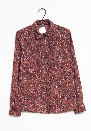 Blouse à manches longues boutonnée avec un motif floral rouge, blanc et bleu, suspendue sur un cintre noir sur fond blanc.