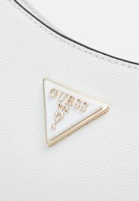 Hvidt tekstureret materiale med et trekantet Guess-logo i guldskrift og et spørgsmålstegn-design.