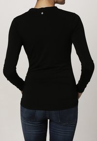 Vrouw in een strak zwart longsleeve shirt en donkerblauwe jeans, gezien vanuit de rug tegen een effen achtergrond.