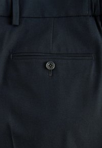 Poche arrière d'un pantalon bleu marine foncé avec une poche passepoil horizontale fermée par un unique bouton noir.