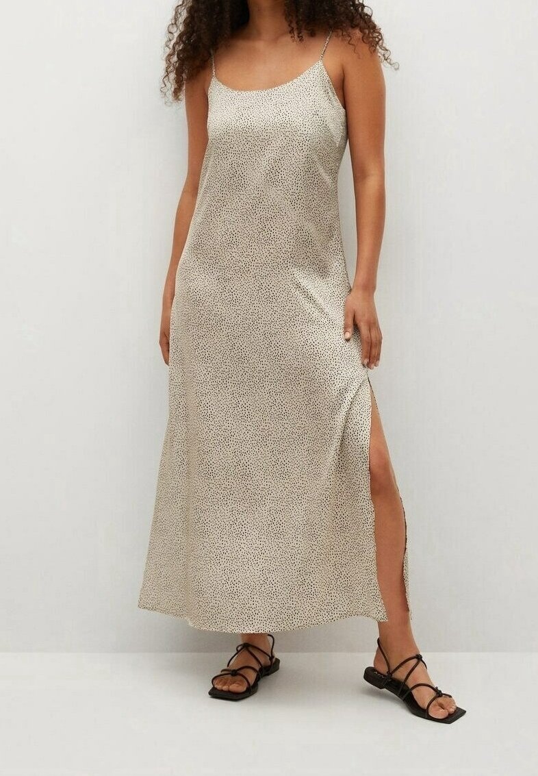 Femme portant une robe maxi beige clair sans manches avec de petits pois noirs et une fente latérale, associée à des sandales plates noires à brides.