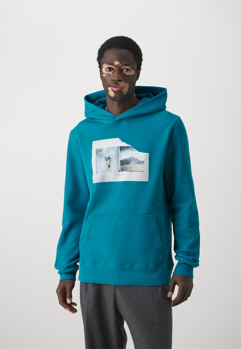Teal hooded sweatshirt features a graphic print of jeans with accents. Includes a front pocket and ribbed cuffs. Pairing with gray pants.