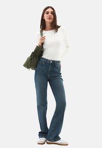 Donna che indossa un top bianco a maniche lunghe, jeans blu a vita alta, sneakers bianche, e tiene una borsa a tracolla verde oliva su uno sfondo neutro.