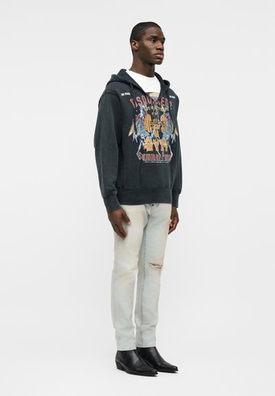 Homme debout de profil face à gauche, portant un sweat à capuche noir avec un imprimé, un T-shirt blanc, un jean clair déchiré et des bottines noires à bout pointu sur fond blanc.