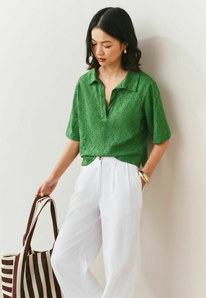 Woman wearing a green eyelet blouse and white pants, holding a striped tote bag, standing against a white wall.