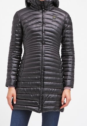 Kobieta w długiej czarnej pikowanej kurtce puchowej z kapturem, z zamkiem z przodu i bocznymi kieszeniami, nosząca niebieskie jeansy.