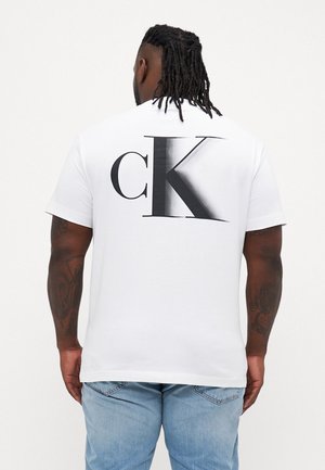 Wit katoenen T-shirt met korte mouwen, met een groot zwart cK-logo op de achterkant. De stof heeft een gladde textuur. Draagt lichtblauwe spijkerbroek.