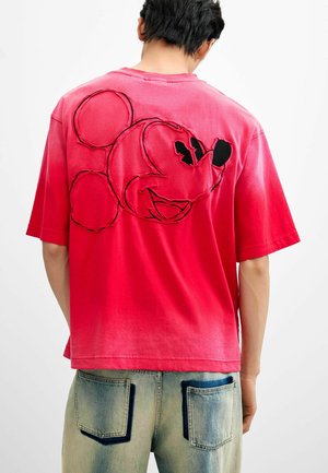 Person trägt ein rotes T-Shirt mit einem umrandeten Mickey-Mouse-Gesichtsdesign auf dem Rücken, kombiniert mit hellblauen Denim-Jeans mit dunklem Taschensaum.