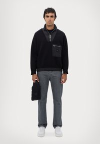 Felpa nera con zip e colletto a contrasto, tasca con zip, abbinata a jeans grigi e sneakers bianche, mentre tiene in mano una piccola borsa nera.