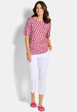 Femme souriante, portant un haut à manches courtes rose à motifs, un pantalon blanc raccourci et des sandales roses tissées, se tenant debout avec une main dans la poche.