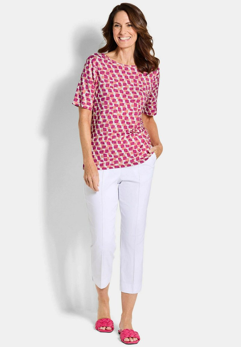 Femme souriante, portant un haut à manches courtes rose à motifs, un pantalon blanc raccourci et des sandales roses tissées, se tenant debout avec une main dans la poche.